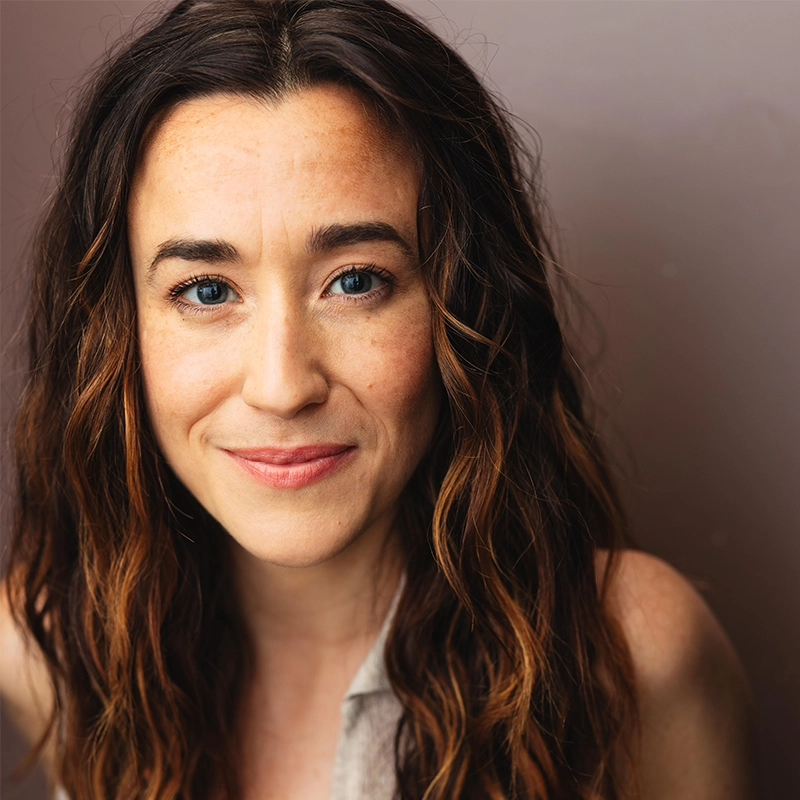 Jessie Shelton, with long wavy brown hair and blue eyes, smiles gently at the camera. She has fair skin with freckles and wears a sleeveless light-colored top. The soft mauve background and warm, natural lighting complete the portrait.