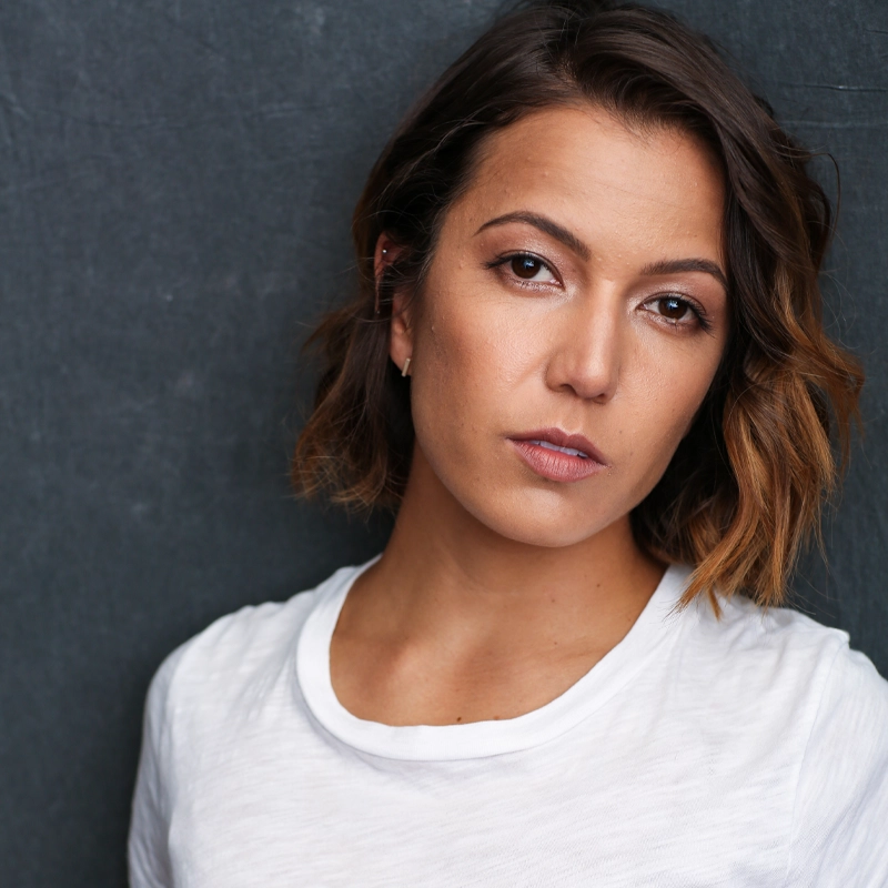 Morgan Marcell, with short, wavy brown hair and light makeup, looks seriously at the camera. She wears a plain white t-shirt and stands against a dark, textured background, her expression neutral with a slight tilt to her head.