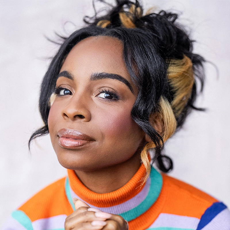 Jessie Shelton, with dark skin and highlighted black hair styled in loose waves, gazes confidently at the camera. She wears natural makeup and a bright orange turtleneck sweater with blue and lavender stripes against a softly blurred background.