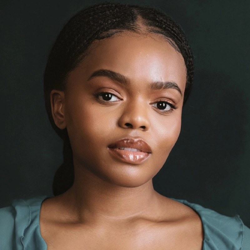 Sandra Okuboyejo, with long, dark hair braided back, gazes forward. Her smooth skin and glossy lips complement a green top against the muted dark background, highlighting her serene expression.