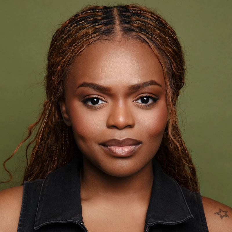 A woman with long, brown braided hair and a star tattoo on her shoulder faces forward, wearing a sleeveless black top. She has smooth skin, neutral makeup, and a calm expression. The background is plain olive green, making Sandra Okuboyejo the focal point of the portrait.