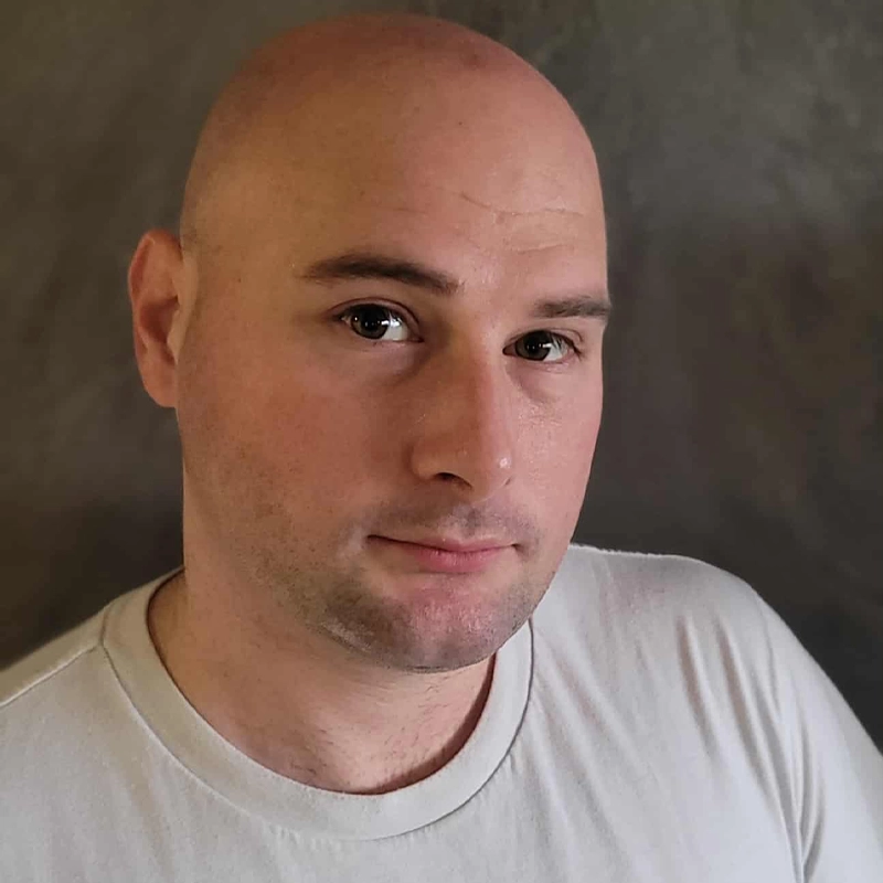 Eric Scherer, a bald man with light skin and brown eyes, is wearing a light-colored T-shirt. He is looking slightly to the side with a neutral expression against a plain, grayish background.