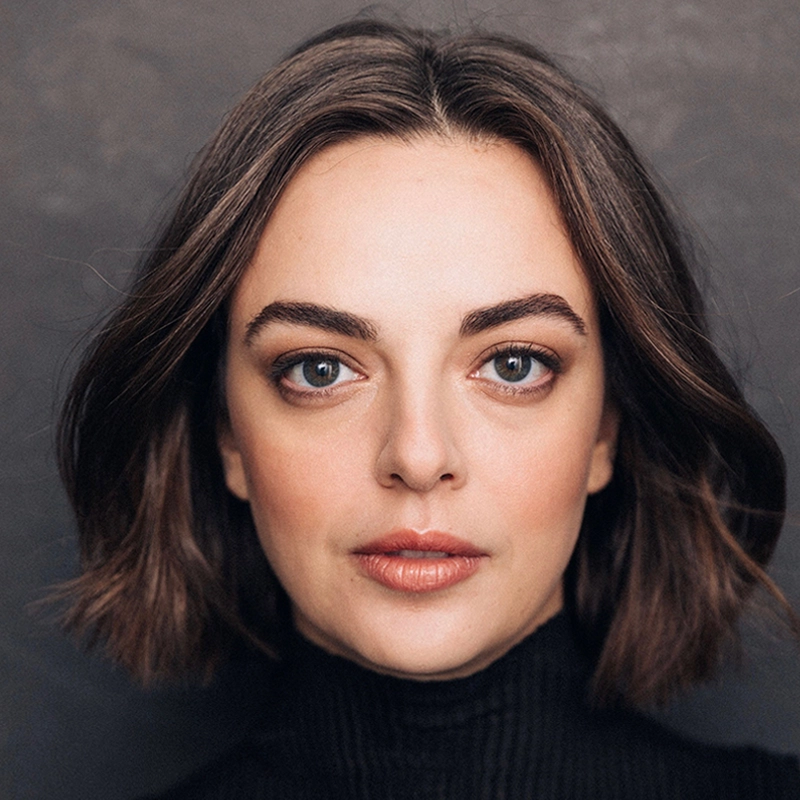 Talia Suskauer, a woman with fair skin, chin-length brown hair, and light eyes, looks directly at the camera. She wears a black turtleneck sweater and subtle makeup. The softly blurred grayish background emphasizes her face.