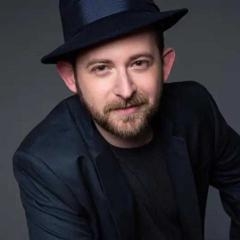A man with a short brown beard and mustache, wearing a black fedora hat, black blazer, and black shirt, poses against a dark gray background. He has a slight smile and is looking directly at the camera.