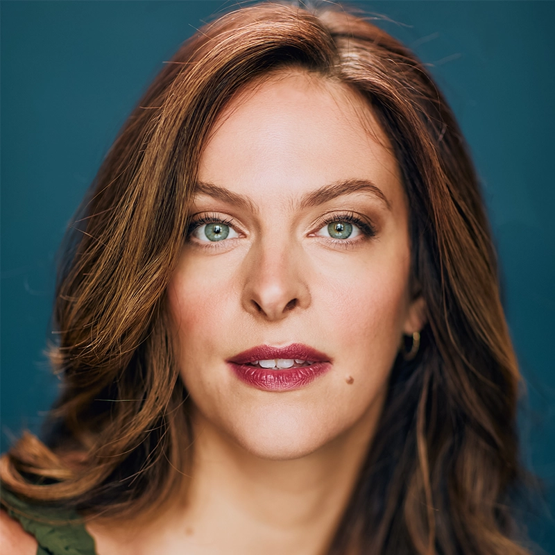 A woman with light skin, long wavy brown hair, and green eyes looks directly at the camera. She is wearing dark red lipstick, light makeup, and a green top—a portrait inspired by the distinctive style of Christian Probst. The background is a solid teal color.