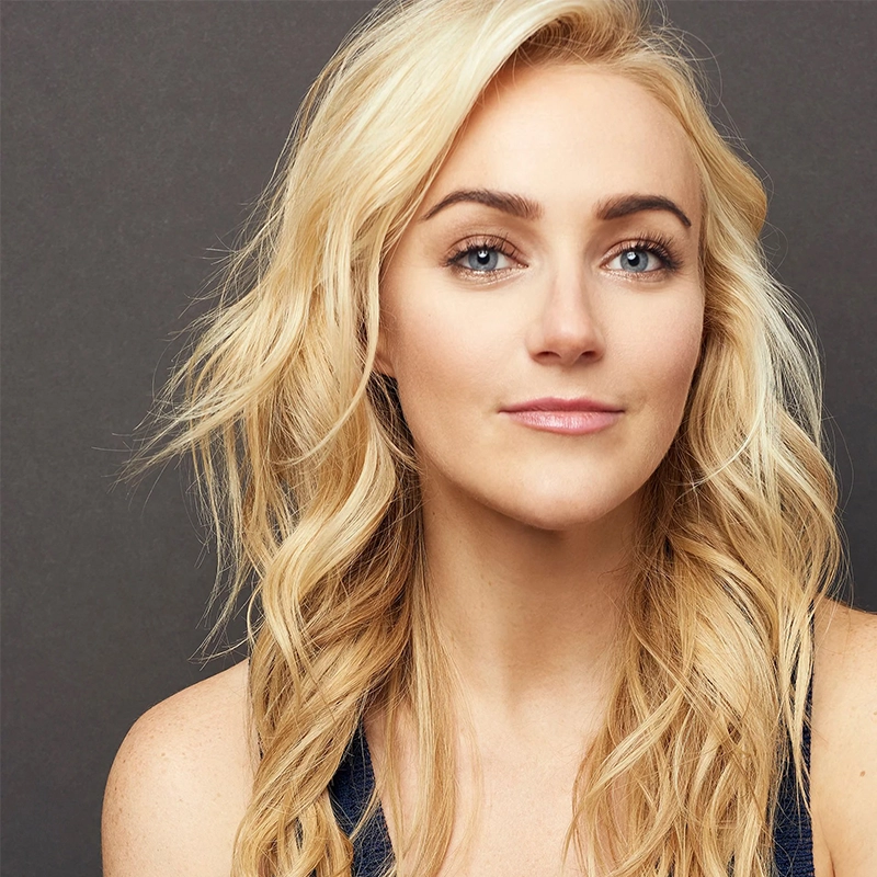 A woman with long, wavy blonde hair and light skin poses in front of a dark gray background. Channeling Jordan Fisher’s effortless style, she has blue eyes, subtle makeup, a slight smile, and wears a dark sleeveless top with her head gently tilted right.