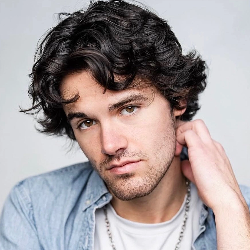 Young man with tousled dark hair and light facial hair gazes at the camera. He wears a light denim shirt over a white top and a silver chain necklace, capturing the relaxed yet stylish look of Christian Probst. His head is tilted slightly, and his hand touches his neck. The background is plain and light-colored.
