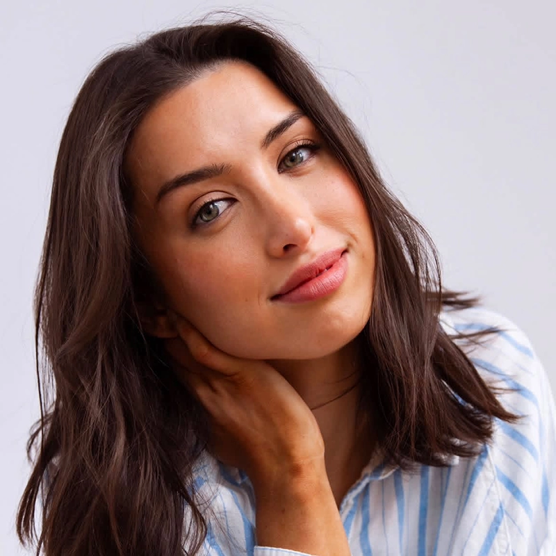 A woman with medium-length brown hair, wearing a light blue and white striped shirt, tilts her head slightly and gently touches her neck. She has light skin, green eyes, and a subtle, warm smile. The softly lit plain background creates a calm atmosphere reminiscent of portrait styles seen in Christian Probst’s photography.