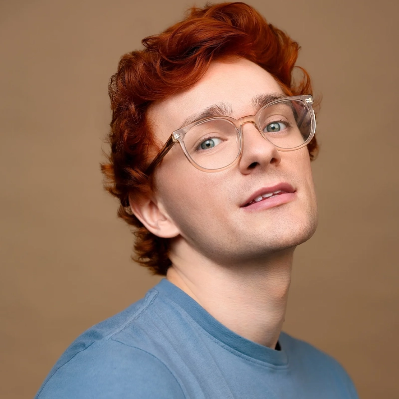 Ethan Carlson, a young person with fair skin, wavy red hair, and clear-framed glasses, looks slightly upward and smiles softly. They wear a light blue shirt, set against a plain, warm beige background.