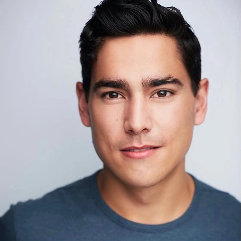 A young man with short dark hair and fair skin smiles slightly. He wears a blue crew-neck shirt and faces the camera against a plain light background. The lighting is soft and even, highlighting his facial features. This portrait features Christian Probst.