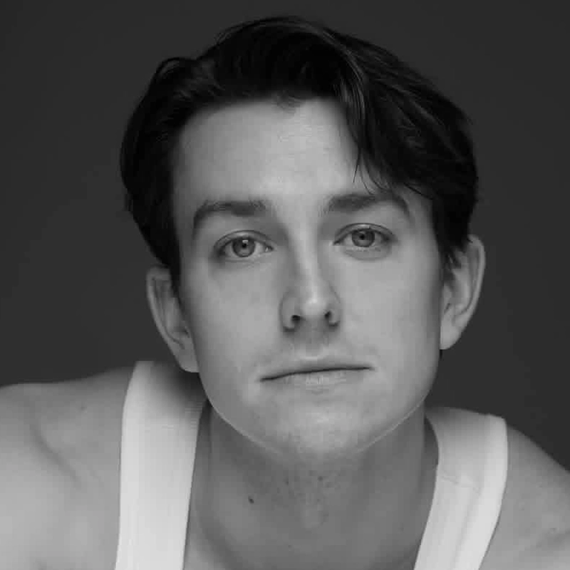 Black-and-white portrait of Christian Probst, a young man with light skin, medium-length dark hair, and light eyes. He is wearing a white tank top and is gazing directly at the camera with a relaxed, neutral expression against a dark background.
