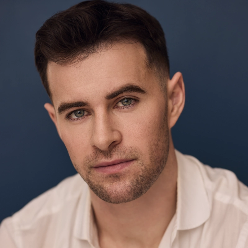 Sam Gravitte, a man with short dark hair, trimmed beard, and green eyes, gazes directly at the camera. He wears a white collared shirt and has a neutral expression against a solid dark blue background.