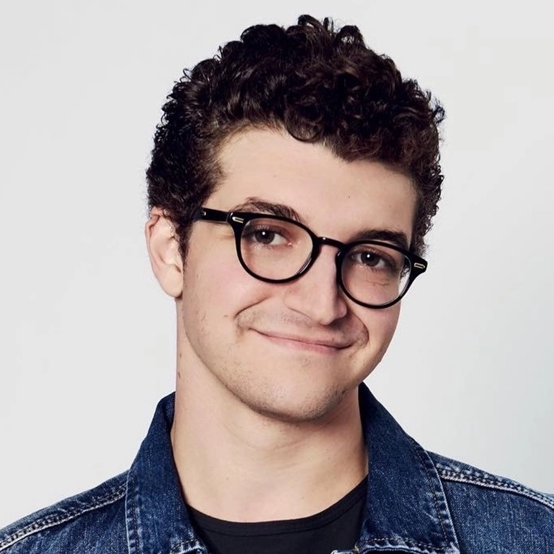 Jared Goldsmith, a young man with short curly brown hair, wearing round black glasses, a blue denim jacket, and a black shirt, smiles slightly while looking at the camera against a plain light background.