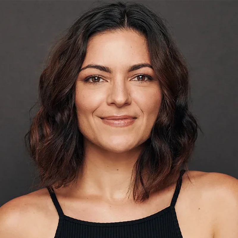 Jennifer DiNoia, with wavy, shoulder-length dark brown hair, smiles gently. She wears a sleeveless black top and stands against a dark, plain background. Her expression is warm and confident.