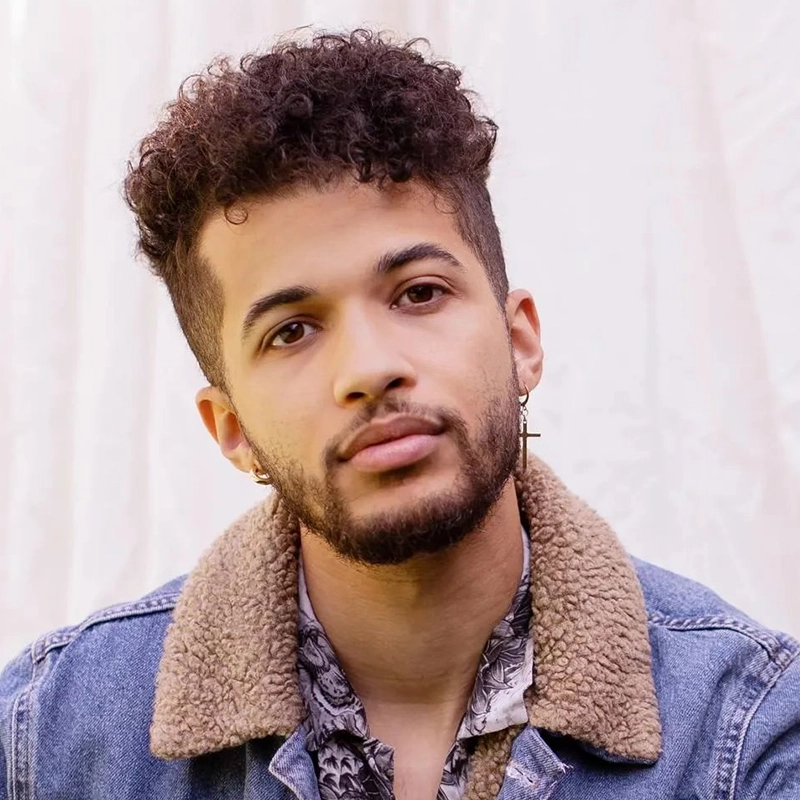 A young man with short curly hair and a trimmed beard, resembling Jordan Fisher, looks at the camera with a neutral expression. He wears a denim jacket with a tan sherpa collar, patterned shirt, and cross-shaped earrings against a blurred fabric background.
