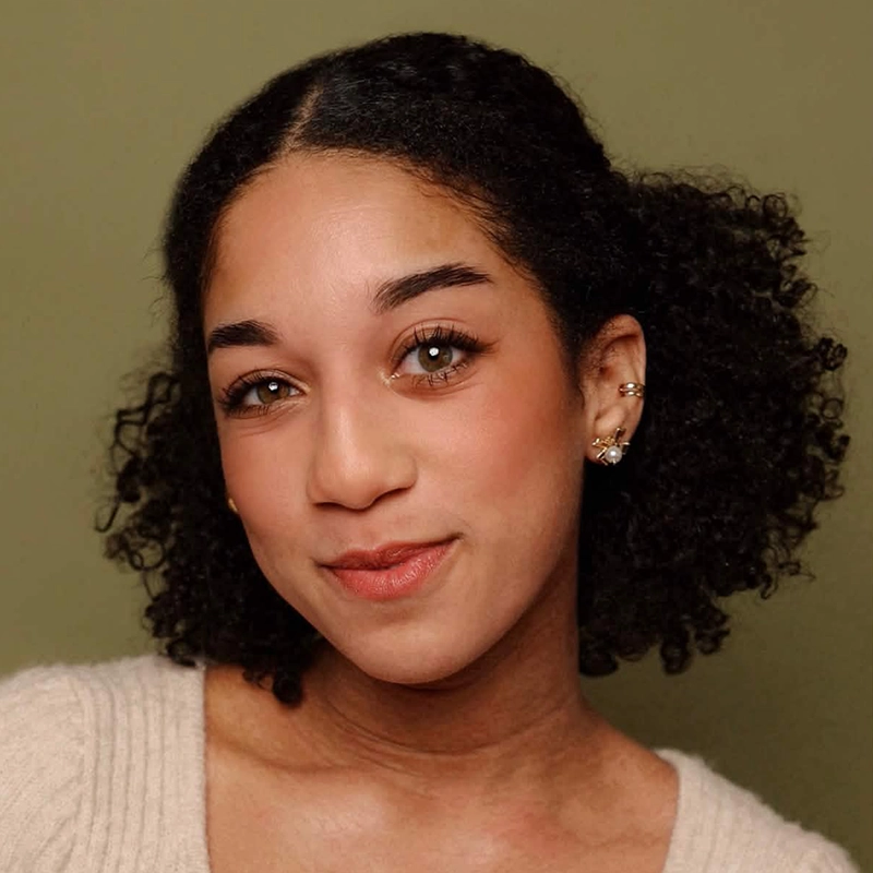 A woman with medium skin, curly dark hair, and brown eyes smiles softly. She wears a cream knit top and multiple earrings. The background is olive green. Her makeup is natural, with subtle blush and lipstick—captured in the signature, expressive portrait style of Christian Probst.