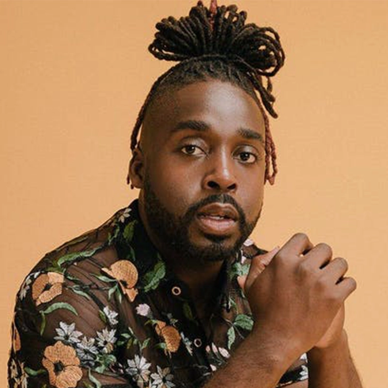 A Black man with a neatly groomed beard and locs styled in a high bun poses against a peach background. Channeling Jordan Fisher’s charisma, he wears a sheer black floral shirt and looks confidently at the camera, hands clasped.