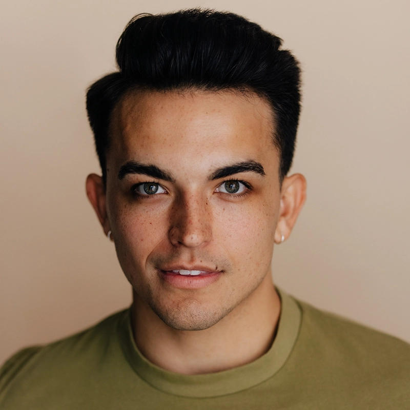 A young man with short dark hair, wearing a green shirt, looks directly at the camera. He has light skin, green eyes, groomed eyebrows, and a slight smile. The background is plain beige, and he wears a small hoop earring in each ear. This portrait features Christian Probst.