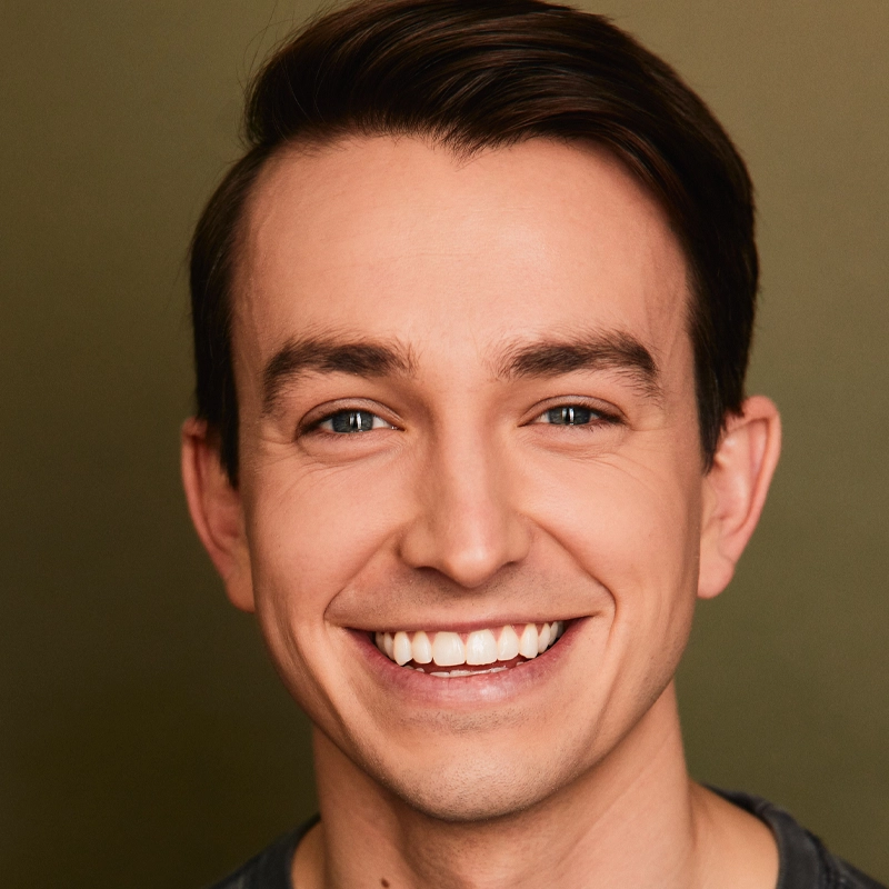 Christian Probst, a young man with fair skin, short dark brown hair, and blue eyes, smiles broadly, showing his teeth. He wears a dark shirt and stands against a plain olive green background with even lighting highlighting his features.