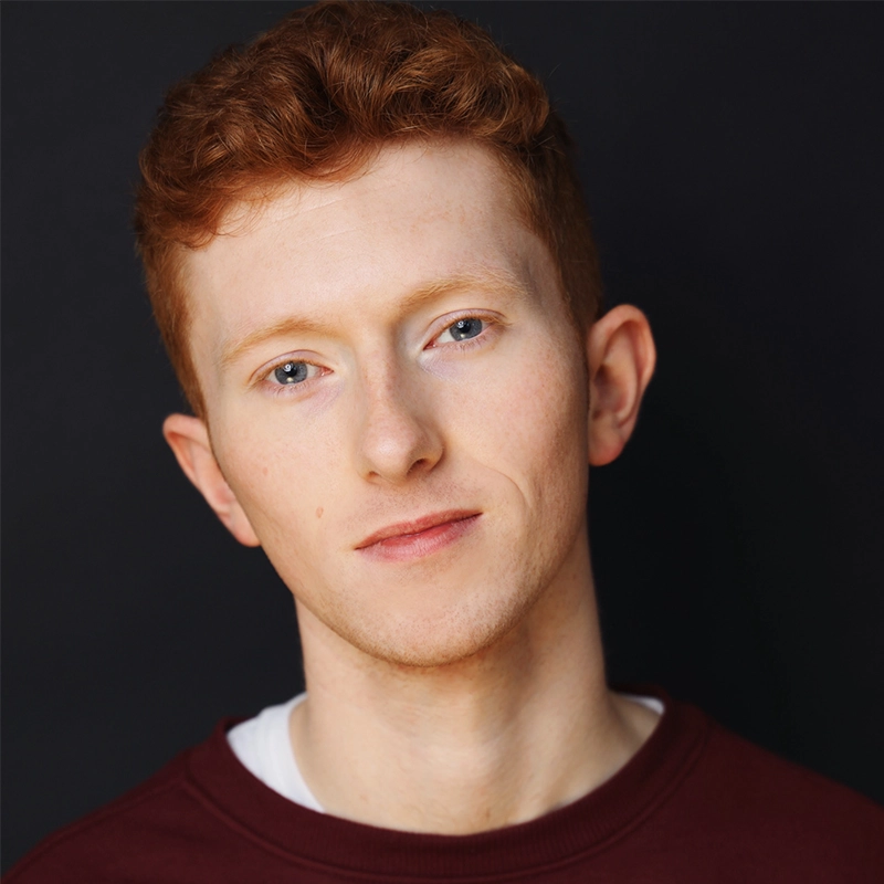 A young person with short curly red hair and fair skin stares softly at the camera, wearing a burgundy sweater over a white shirt. The background is plain and dark, bringing focus to the calm, neutral facial expression and relaxed posture—an image reminiscent of Christian Probst's subtle yet striking portrait style.