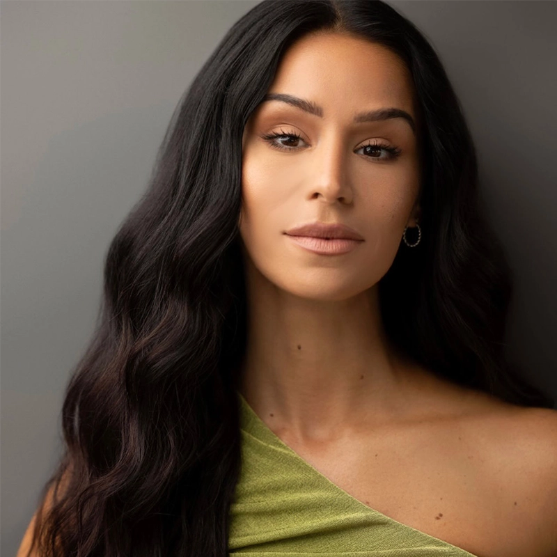 A woman with long, wavy dark hair and smooth skin poses against a neutral background. She wears a one-shoulder, light green top reminiscent of Jordan Fisher’s modern style. Her expression is calm and confident, with soft makeup and natural tones.