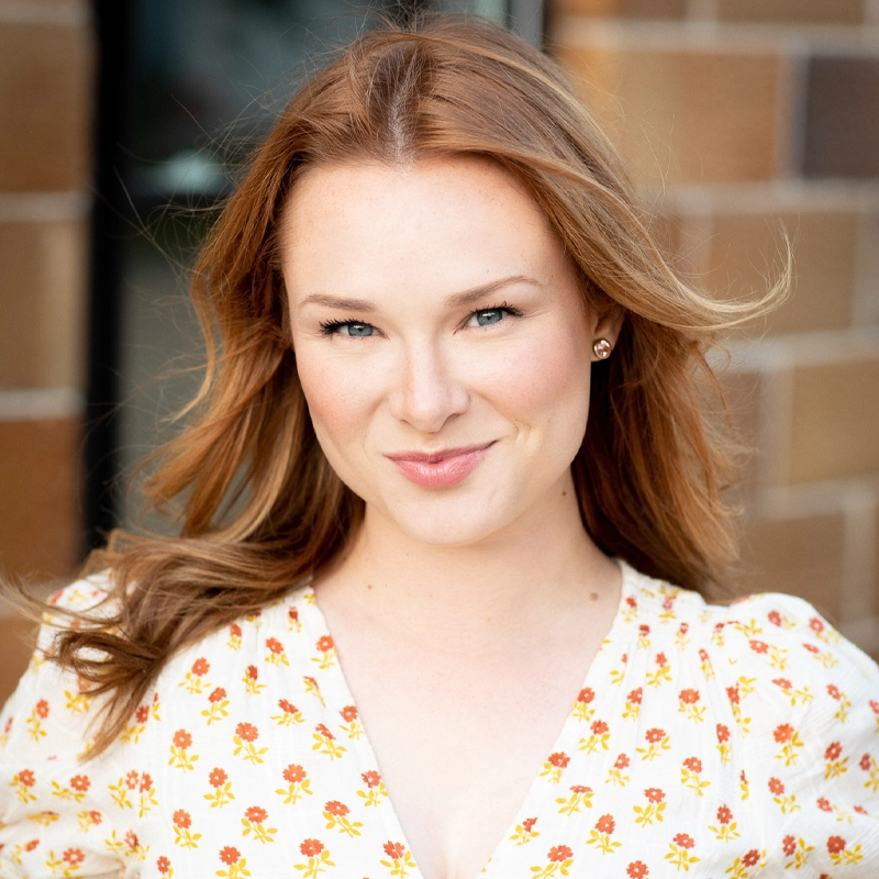 A woman with fair skin and long, wavy, light brown hair smiles confidently. Erin Stapleton wears a cream floral top with orange and yellow flowers. The background is a blurred brick wall. The lighting is natural, highlighting her face and hair.