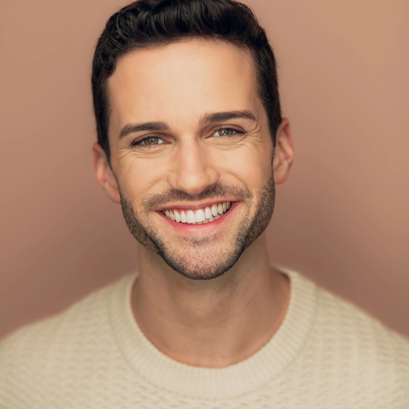 Michael Williams, a young man with short dark hair and a trimmed beard, smiles brightly at the camera. He is wearing a cream-colored textured sweater against a soft, warm beige background, creating a friendly and approachable atmosphere.