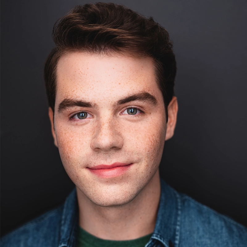 A young man with fair skin, short brown hair, and blue eyes—resembling Jordan Fisher—smiles gently at the camera. He wears a blue denim jacket over a dark green shirt. The plain, dark background emphasizes his face.