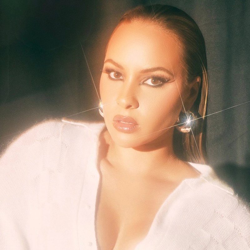 A woman with slicked-back hair wears large hoop earrings and a white, buttoned top. Reminiscent of actress Jasmine Cephas Jones from Hamilton, her bold eyeliner and glossy lips stand out in the glamorous, ethereal glow with soft light flares.