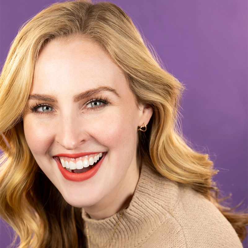 A smiling woman, Catherine DeLuce, with wavy blonde hair wearing a beige sweater and small hoop earrings poses in front of a solid purple background. She is looking toward the camera, showing her teeth and bright red lipstick.