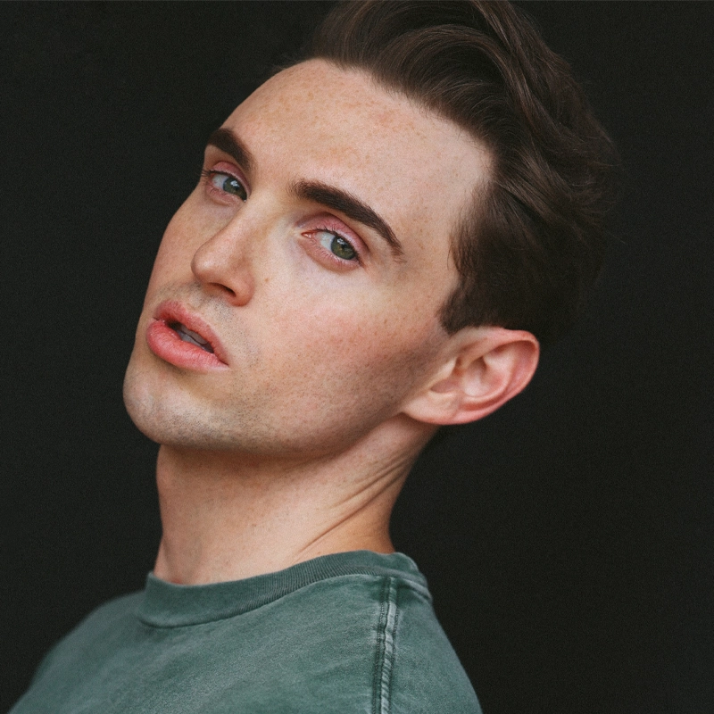 A young adult with fair skin, short brown hair, and blue-green eyes wears a faded green crewneck shirt. Their head is tilted back and turned slightly to the side, with a neutral expression. The background is solid black. This is Joe Hornberger.