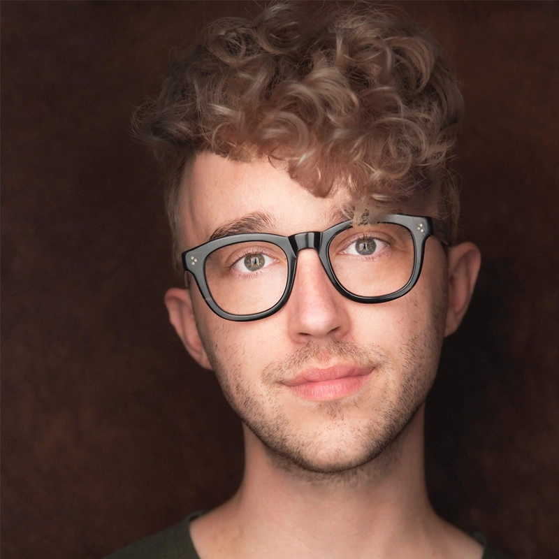 A young person with light skin, curly brown hair, and black-rimmed glasses looks directly at the camera. They have slight stubble and wear a green shirt. The background is softly blurred in a dark, warm tone.