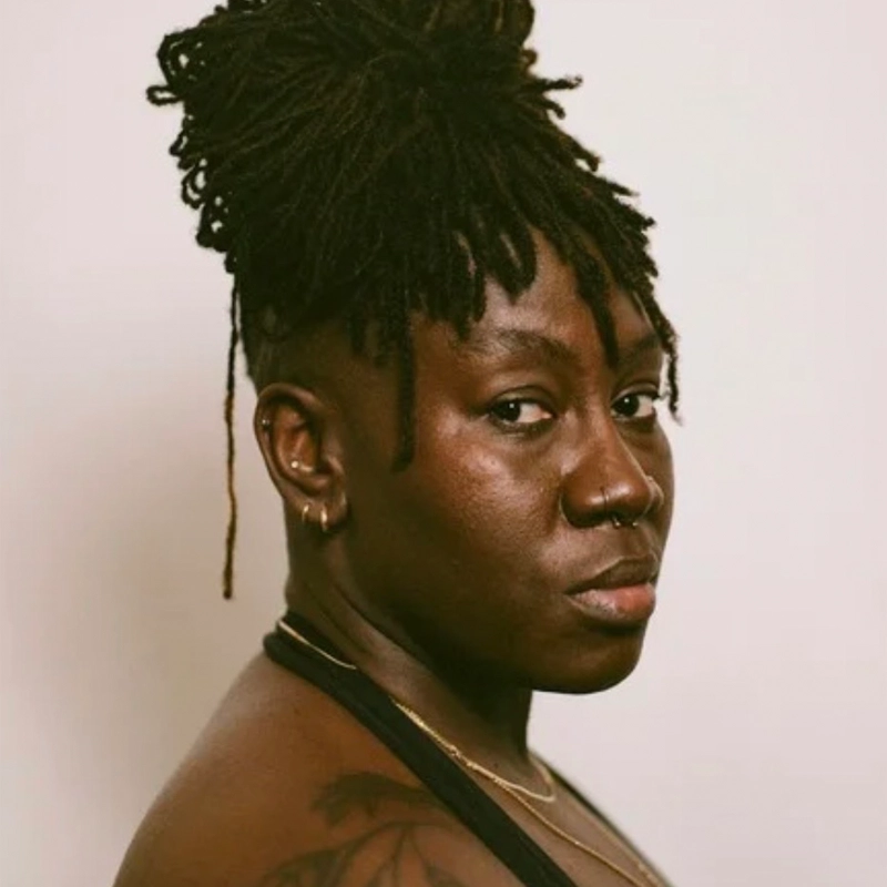 A person with dark skin and long, thick dreadlocks styled in a high bun looks at the camera with a serious expression. They have a nose ring, hoop earrings, and a tattoo on their shoulder. The background is plain and light-colored.