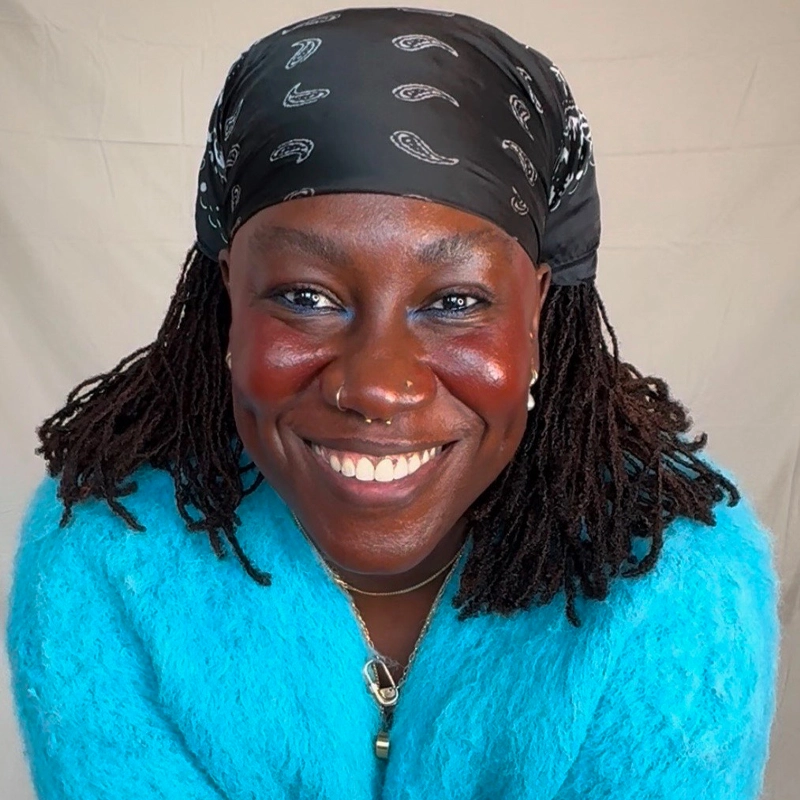A person with dark skin, blue eyeshadow, and red blush smiles warmly. They wear a black bandana with white paisley patterns, long twisted hair, a fuzzy bright blue sweater, layered necklaces, and a nose ring. The background is a neutral beige.