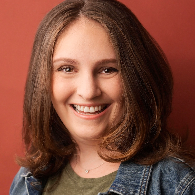 A woman with shoulder-length light brown hair, smiling, faces the camera. She has fair skin, wears a denim jacket over a green shirt, and a delicate necklace. The background is a solid, warm reddish color. This is Dara Paige Bloomfield.