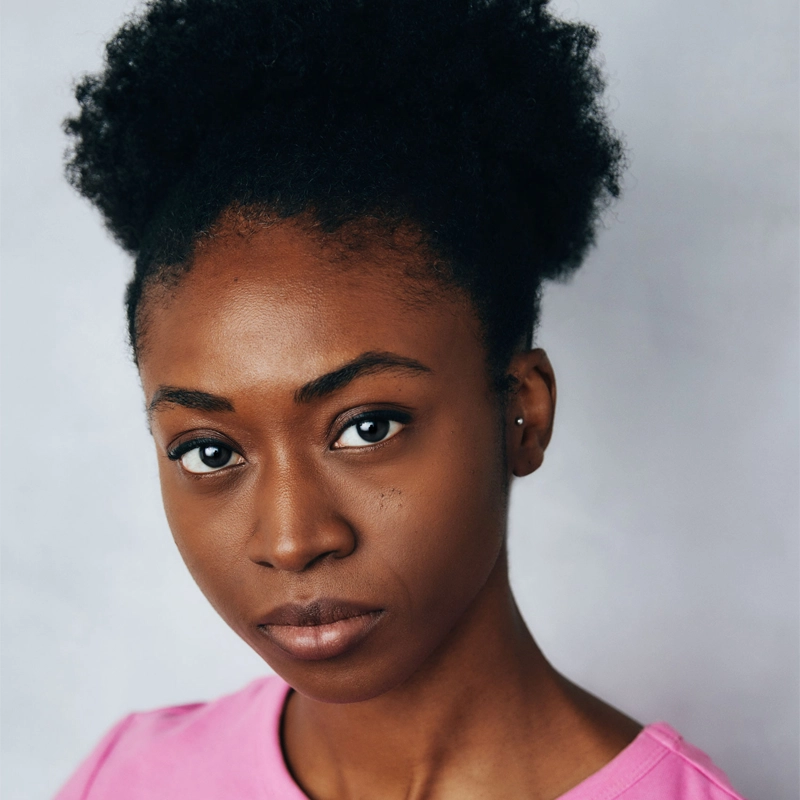 A young Black woman with natural hair in a curly puff looks directly at the camera with a neutral expression. She is wearing a pink top and small earring. The background is plain and light, keeping the focus on her face.