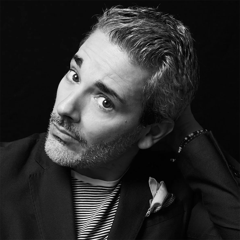 Black and white portrait of Eric Yves Garcia with short, wavy salt-and-pepper hair and a trimmed beard, wearing a striped shirt under a dark blazer. He looks upward, tilting his head with one hand behind his neck against a dark background.