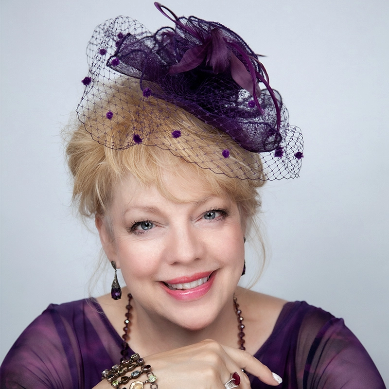 A smiling woman with light blonde hair wears a sheer purple fascinator hat with netting and feathers, matching purple earrings, a purple sheer top, and several bracelets. She poses with her hand under her chin against a light background.