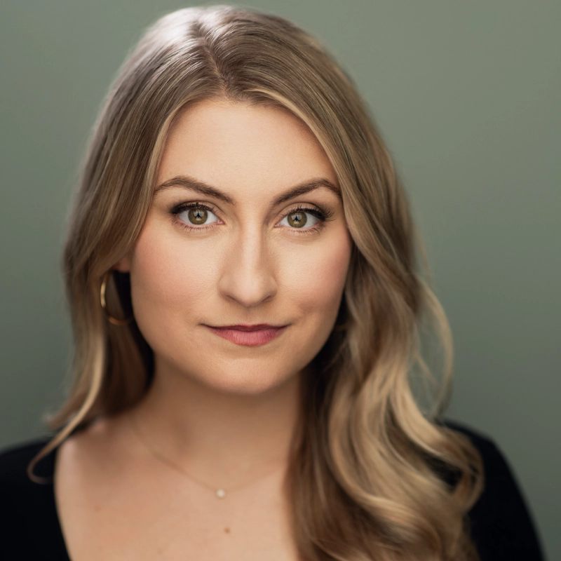 Aashley Morgan, with long wavy blonde hair and hazel eyes, looks directly at the camera, wearing a black top, gold hoop earring, and a delicate necklace against a muted green background with soft lighting.