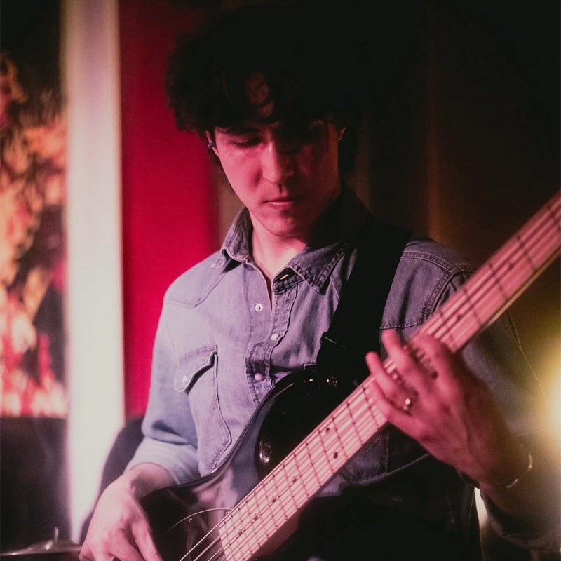 A young man with dark hair wearing a denim shirt plays an electric bass guitar. Caleb Pringle is focused on the instrument, with soft, moody lighting and a blurred, colorful background suggesting a live music performance or intimate setting.