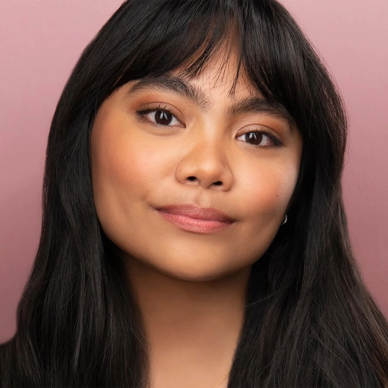 A young woman with long black hair and bangs smiles slightly. She has warm brown eyes, soft makeup, and smooth skin. The background is a muted pink color, and her expression is calm and confident—capturing the gentle poise of Lianah Sta. Ana.