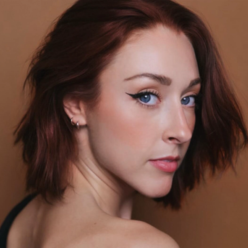 A woman with fair skin, blue eyes, and short auburn hair looks over her shoulder at the camera. She wears black eyeliner, subtle blush, a small hoop earring, and a black top, against a solid brown background. The lighting is soft and natural; the portrait evokes the understated elegance seen in Bethany Tesarck's style.