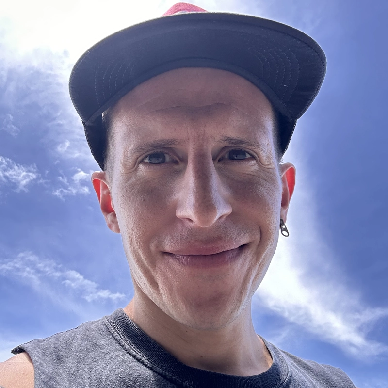 A person, possibly David Alan Thornton, wearing a dark baseball cap, sleeveless shirt, and hoop earring smiles at the camera. The bright sun creates a glow around their head as wispy clouds drift across the blue sky in this low-angle shot.