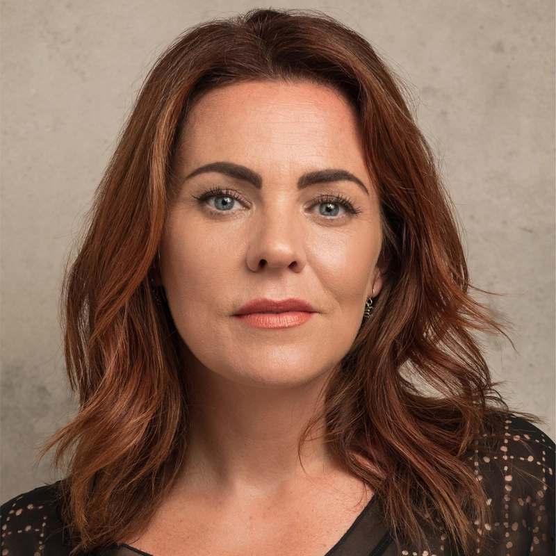 Rachel Tucker, a woman with fair skin and shoulder-length auburn hair, poses against a neutral, textured background. She has blue eyes, lightly arched brows, and wears subtle makeup with natural lipstick and a black top with sheer dotted sleeves.