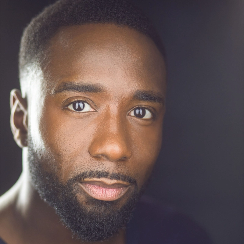A close-up portrait of John Edwards, a man with short black hair and a full beard, looking directly at the camera. He has dark brown eyes, smooth skin, and wears a neutral expression. The dark background softly highlights his face.