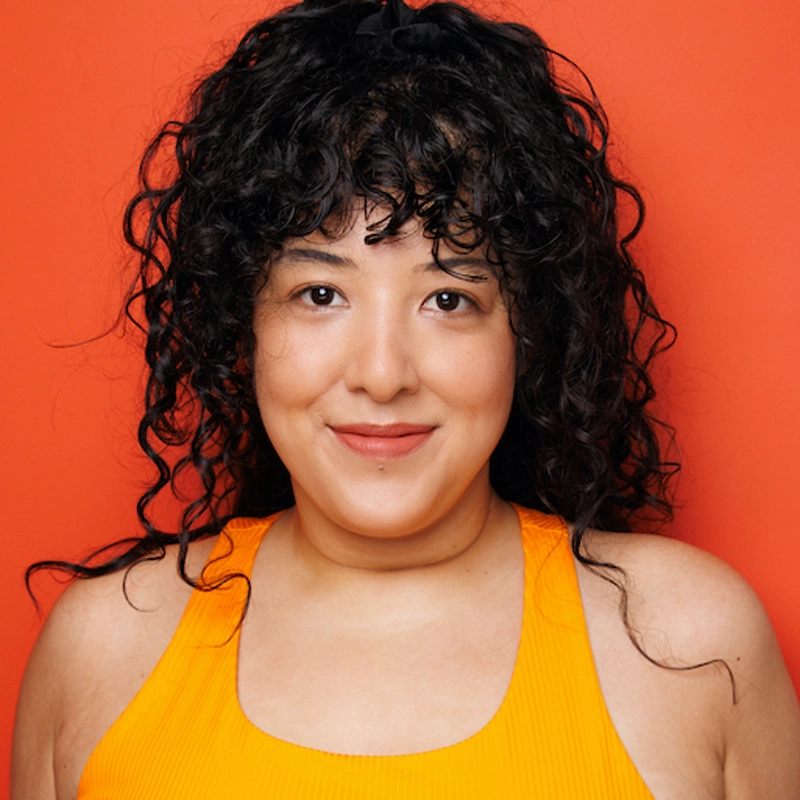 Alina Fontanilla, with curly dark hair and bangs, smiles softly at the camera. She wears a bright yellow sleeveless top against an orange background, creating a vibrant, warm color contrast. Her expression is friendly and relaxed.
