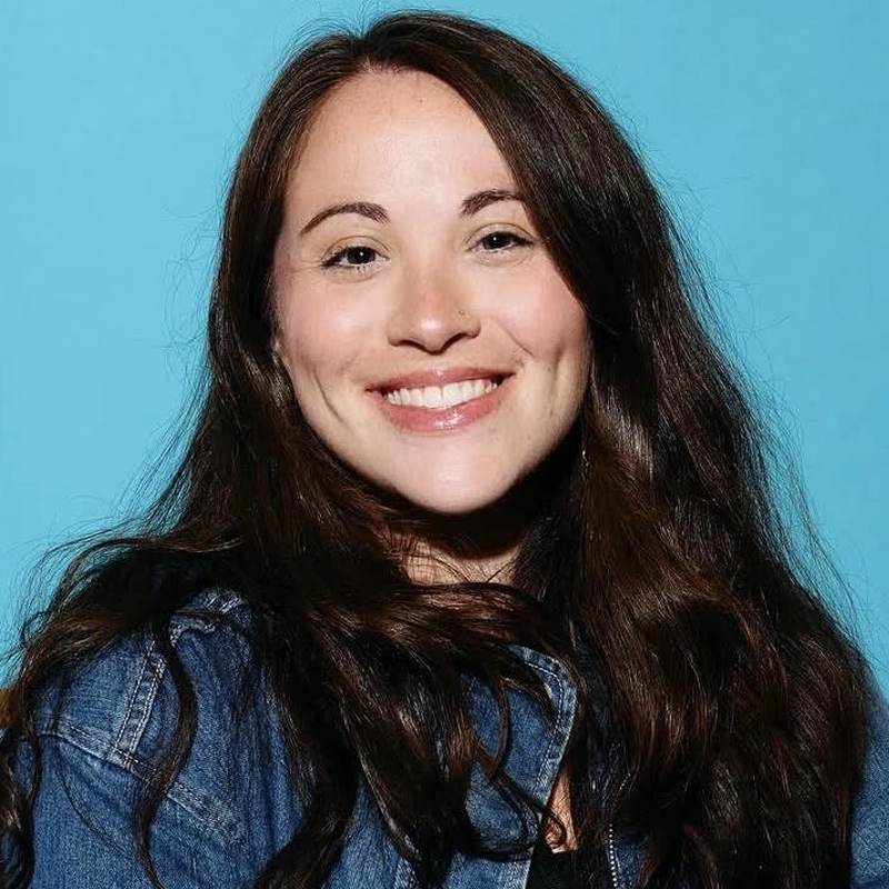 Molly Heller, a woman with long, wavy brown hair, smiles at the camera. She wears a blue denim jacket over a black top. The background is solid light blue. Her expression is friendly and she has dimples on her cheeks.