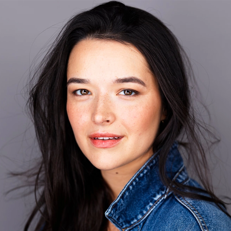 Kailee Graham, a woman with long dark hair parted slightly off-center, wears a denim jacket and looks at the camera. She has light skin, brown eyes, natural makeup, and a slight smile. The background is a plain grey.
