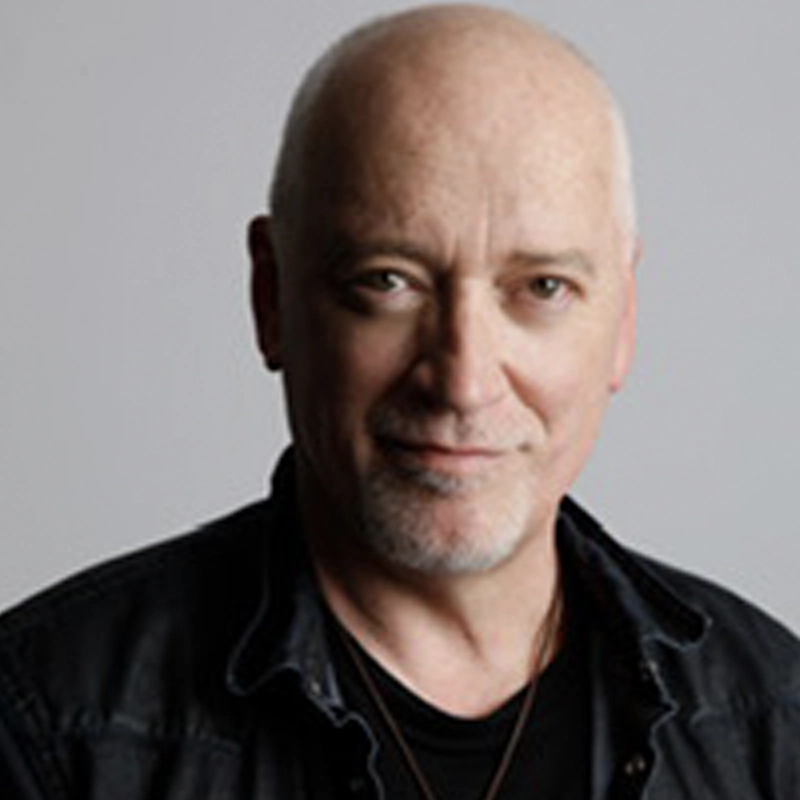 A middle-aged, bald man with light skin and blue eyes smiles gently. He wears a black shirt and a dark jacket against a plain, light gray background. The lighting is soft, highlighting his friendly expression.