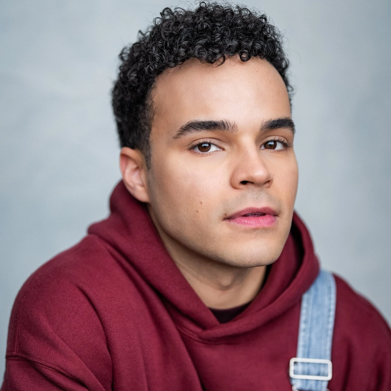Chris Medlin, a young person with short, curly black hair and medium skin tone, wears a maroon hoodie layered under blue denim overalls. They gaze calmly at the camera against a soft, light gray background with even lighting highlighting their features.
